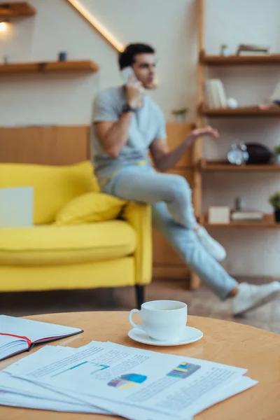 Foco seletivo de mesa de café com papéis, xícara de café e notebook com freelancer falando no smartphone no sofá — Fotografia de Stock