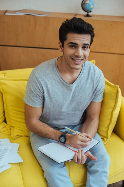 High angle view of freelancer writing in notebook, smiling and looking at camera on sofa — Stock Photo