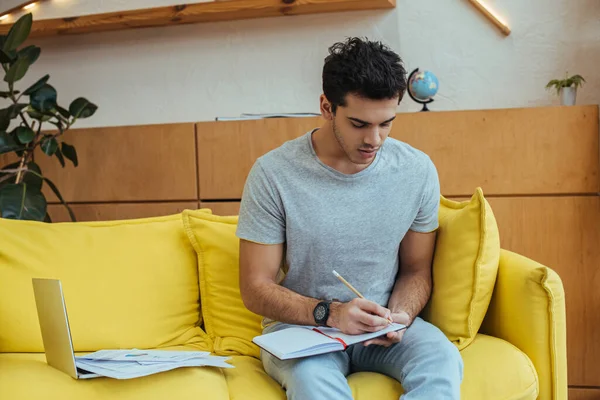Freiberuflerin schreibt in Notizbuch neben Laptop mit Papieren auf Sofa im Wohnzimmer — Stockfoto