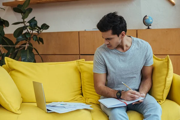 Freelancer con notebook mirando portátil con papeles en sofá en sala de estar - foto de stock