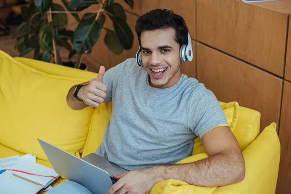 Hochwinkelaufnahme eines Freelancers mit Kopfhörer und Laptop, der lächelnd in die Kamera blickt und sich wie ein Schild auf dem Sofa zeigt — Stockfoto