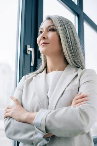 Confiada mujer de negocios asiática con pelo gris y brazos cruzados en la oficina - foto de stock