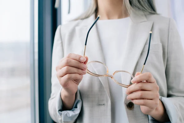Обрізаний вид виконавчої бізнес-леді в сірому костюмі, що тримає окуляри в офісі — стокове фото