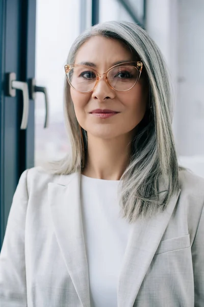 Atractiva mujer de negocios asiática con pelo gris en traje gris y gafas en la oficina - foto de stock