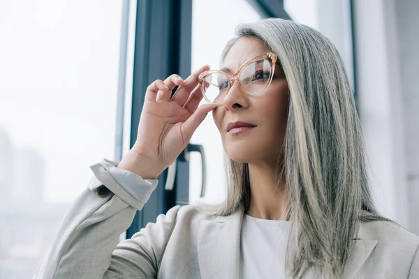 Femme d'affaires asiatique réfléchie avec des cheveux gris en costume gris et des lunettes au bureau — Photo de stock