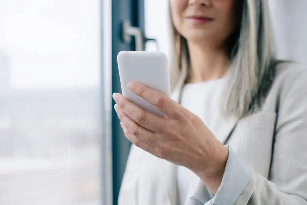 Обрізаний вид бізнес-леді з сірим волоссям, використовуючи смартфон в офісі — стокове фото