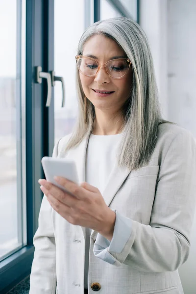 Дорослий усміхнений азіатський бізнесмен в окулярах з сірим волоссям, використовуючи смартфон в офісі — стокове фото