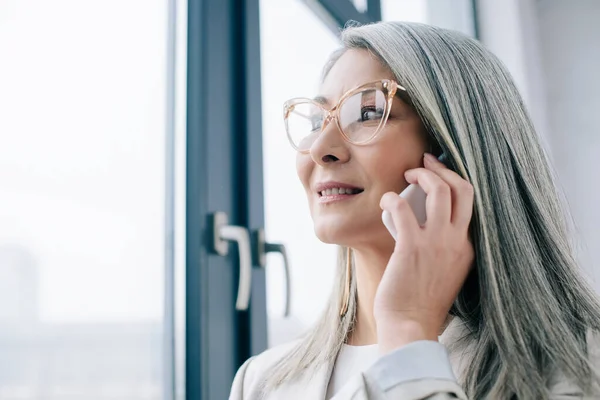 Professional asian businesswoman in eyeglasses with grey hair talking on smartphone in office — Stock Photo