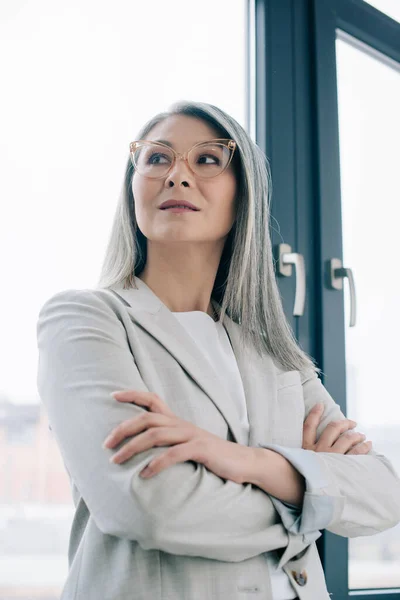 Корпоративная азиатская бизнесвумен со скрещенными руками и седыми волосами, стоящая в сером костюме и очках в офисе — стоковое фото
