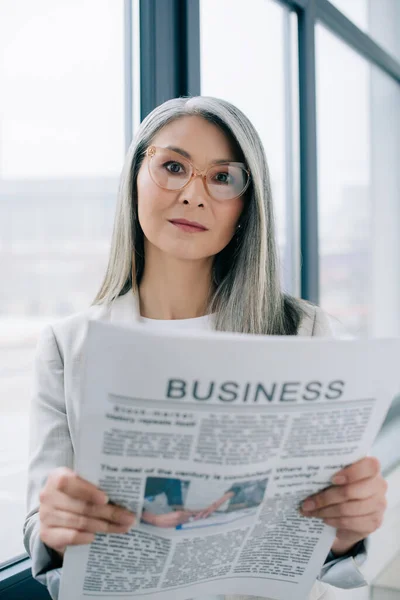 Fiduciosa donna d'affari asiatica in occhiali in possesso di giornale aziendale in ufficio — Foto stock