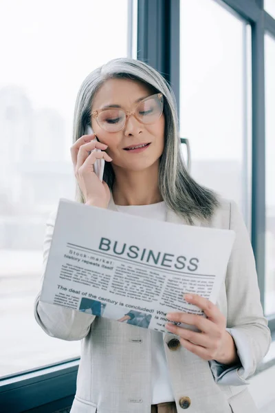 Attraktive asiatische Geschäftsfrau mit Brille spricht auf dem Smartphone, während sie im Büro Wirtschaftszeitung liest — Stockfoto