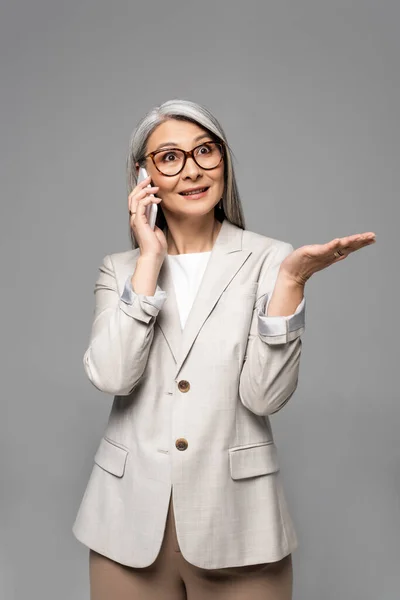 Surprised asian businesswoman in eyeglasses with grey hair talking on smartphone isolated on grey — Stock Photo
