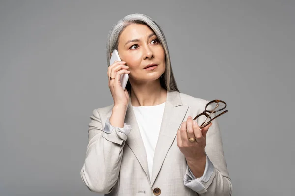 Напружена азіатська бізнес-леді з сірим волоссям розмовляє на смартфоні і тримає окуляри ізольовані на сірому — стокове фото