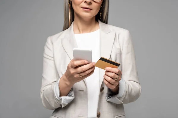 Обрізаний вид бізнес-леді з сірим волоссям покупки в Інтернеті з кредитною карткою і смартфон ізольовані на сірому — стокове фото