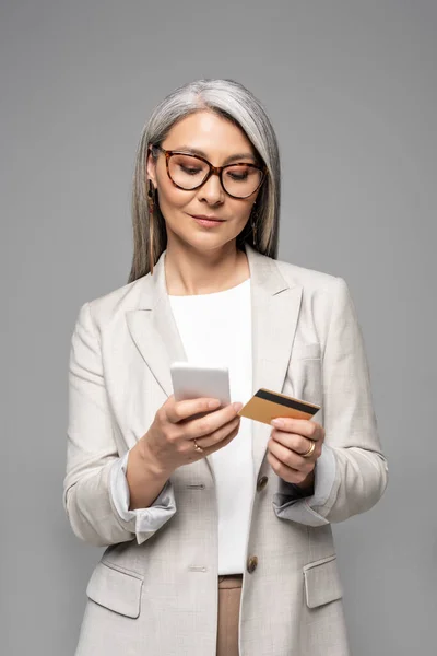 Приваблива азіатська бізнес-леді з сірим волоссям покупки в Інтернеті з кредитною карткою і смартфон ізольовані на сірому — стокове фото
