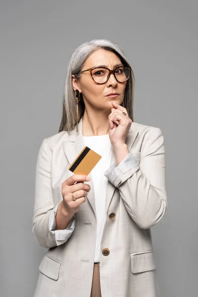 Задумлива азіатська бізнес-леді з сірим волоссям тримає кредитну картку ізольовано на сірому — стокове фото