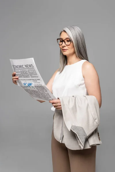 Уверенная азиатская бизнесвумен в очках, читающая деловую газету, изолированную на сером — стоковое фото