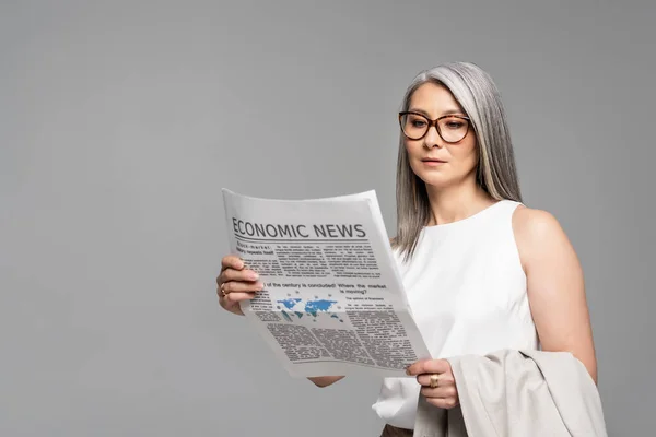 Beautiful asian businesswoman in eyeglasses reading business newspaper isolated on grey — Stock Photo