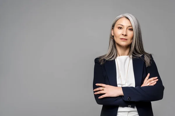 Confiada mujer de negocios asiática con pelo gris y brazos cruzados aislados en gris - foto de stock