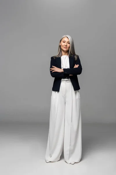 Fiduciosa donna d'affari asiatica con i capelli grigi e braccia incrociate sul grigio — Foto stock