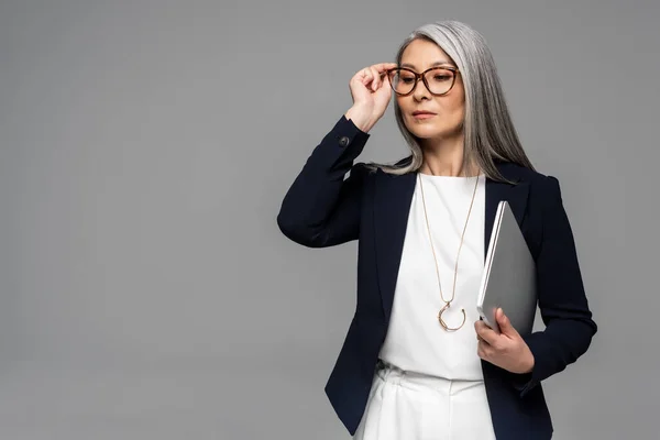 Attraktive selbstbewusste asiatische Geschäftsfrau mit grauen Haaren hält Laptop isoliert auf grau — Stockfoto