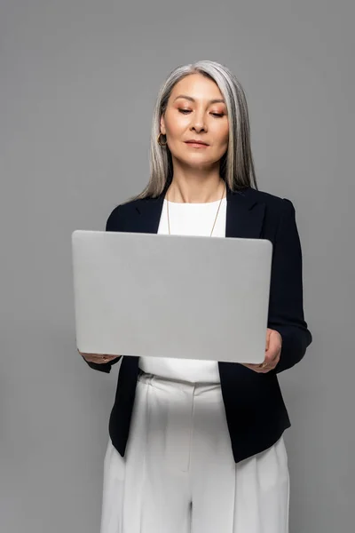 Atraente asiático empresária com cinza cabelo usando laptop isolado no cinza — Fotografia de Stock