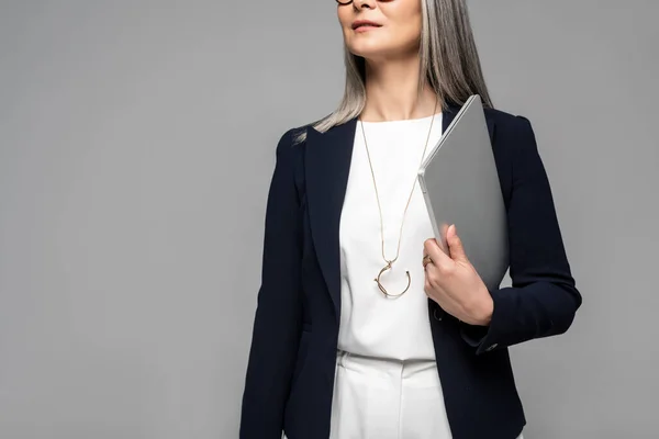 Обрізаний вид бізнес-леді з сірим волоссям тримає ноутбук ізольований на сірому — стокове фото