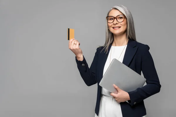 Heureux asiatique femme d'affaires avec gris cheveux shopping en ligne avec carte de crédit et ordinateur portable isolé sur gris — Photo de stock