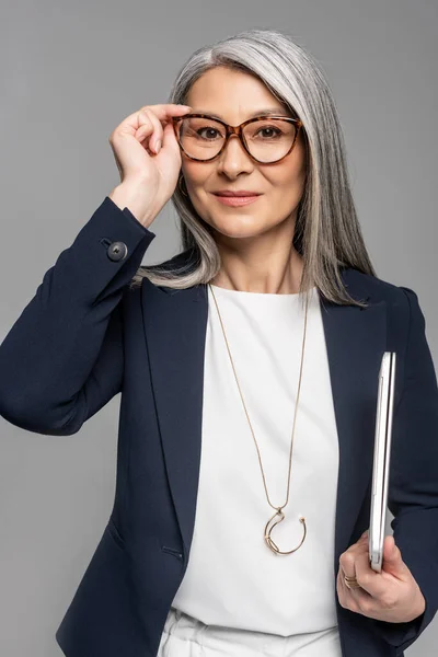 Lächelnde asiatische Geschäftsfrau mit grauen Haaren hält Laptop isoliert auf grau — Stockfoto