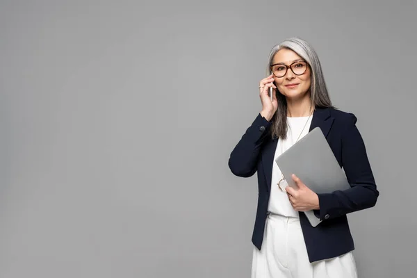 Entreprise asiatique femme d'affaires avec des cheveux gris parler sur smartphone et tenant ordinateur portable isolé sur gris — Photo de stock
