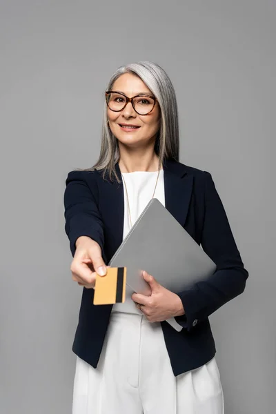Lächelnde asiatische Geschäftsfrau mit grauen Haaren shoppt online mit Kreditkarte und Laptop isoliert auf grau — Stockfoto