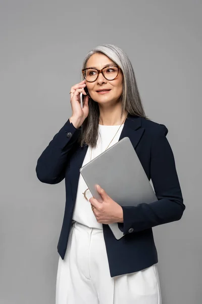 Attractive asian businesswoman with grey hair talking on smartphone and holding laptop isolated on grey — Stock Photo