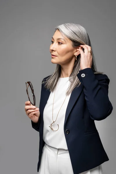 Mujer de negocios asiática con pelo gris sosteniendo gafas aisladas en gris - foto de stock