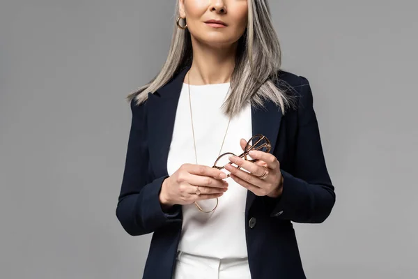 Abgeschnittene Ansicht einer Geschäftsfrau mit grauen Haaren, die eine auf grau isolierte Brille hält — Stockfoto