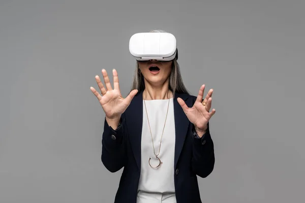 Шокована бізнес-леді жестикулює і використовує гарнітуру віртуальної реальності ізольовано на сірому — стокове фото