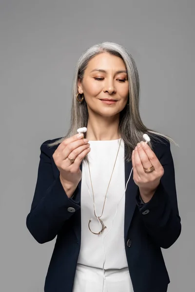 Sonriente asiático mujer de negocios con gris pelo celebración auriculares aislado en gris - foto de stock