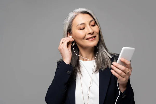 Smiling asian businesswoman with grey hair listening music with earphones and smartphone isolated on grey — Stock Photo