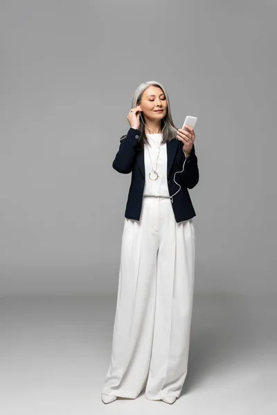 Attractive asian businesswoman with grey hair listening music with earphones and smartphone on grey — Stock Photo