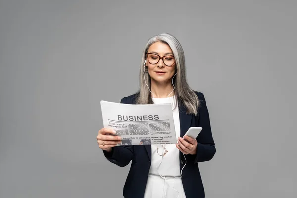 Attractive asian businesswoman in eyeglasses listening music with earphones and smartphone while reading business newspaper isolated on grey — Stock Photo