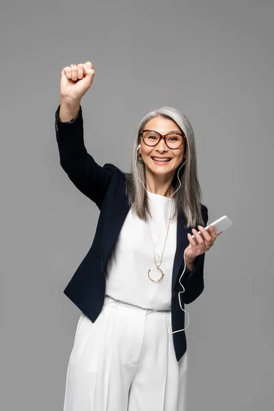 Excité asiatique femme d'affaires avec gris cheveux écoute de la musique avec écouteurs et smartphone isolé sur gris — Photo de stock