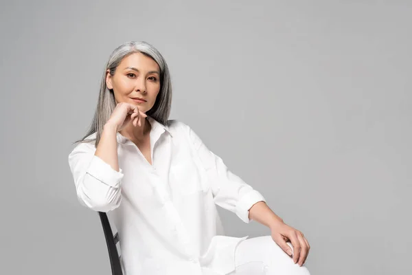 Серйозна азіатська жінка з сірим волоссям сидить на стільці ізольовано на сірому — стокове фото