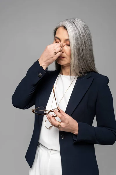 Tired asian businesswoman with grey hair holding eyeglasses isolated on grey — Stock Photo