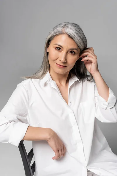 Задумлива азіатська жінка з сірим волоссям сидить на стільці ізольовано на сірому — стокове фото