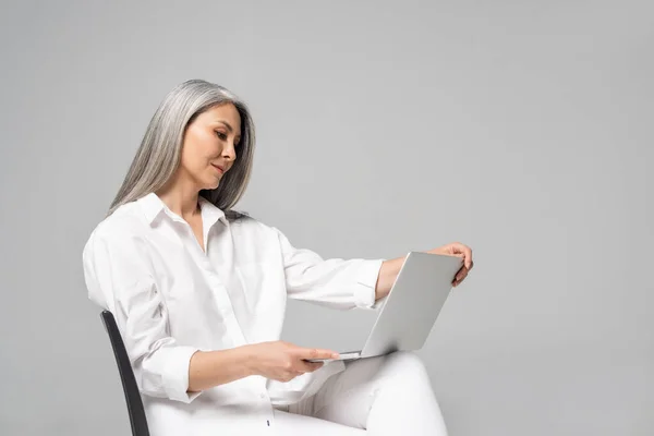Доросла азіатка з сірим волоссям сидить на стільці і використовує ноутбук ізольовано на сірому — стокове фото
