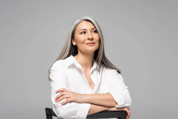 Красива мрійлива азіатська жінка з сірим волоссям сидить на стільці ізольована на сірому — стокове фото