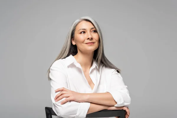 Bela sonhador asiático mulher com cinza cabelo sentado no cadeira isolado no cinza — Fotografia de Stock