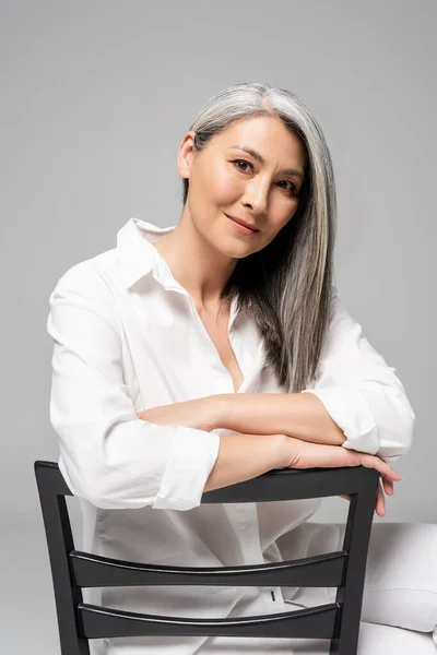 Усміхнена азіатська жінка з сірим волоссям сидить на стільці ізольовано на сірому — стокове фото