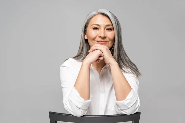 Beautiful positive asian woman with grey hair sitting on chair isolated on grey — Stock Photo