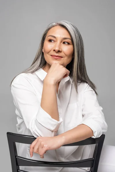 Щаслива красива азіатська жінка з сірим волоссям сидить на стільці ізольована на сірому — стокове фото