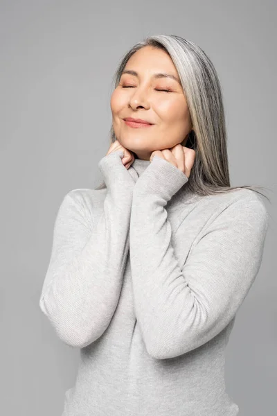 Smiling asian woman in turtleneck with grey hair and closed eyes isolated on grey — Stock Photo